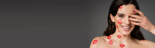 Happy shirtless woman with red kiss prints on face and body covering eye with hand isolated on grey, banner — Stock Photo