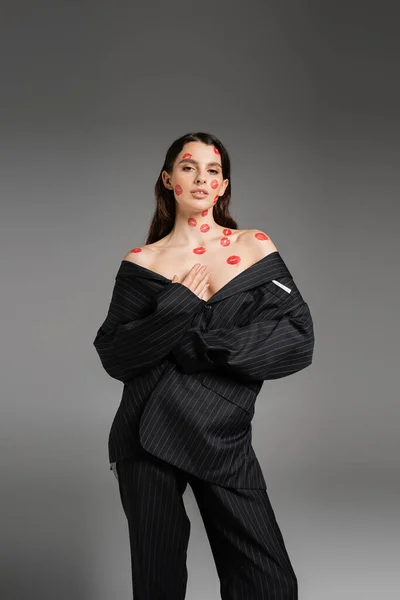 Young woman with red lipstick marks on naked shoulders and face posing in oversize suit isolated on grey — Stock Photo