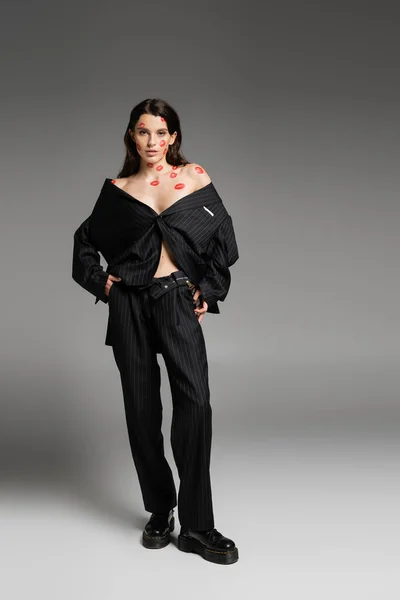 Full length of brunette woman with red kiss prints wearing black oversize suit and looking at camera on grey — Stock Photo