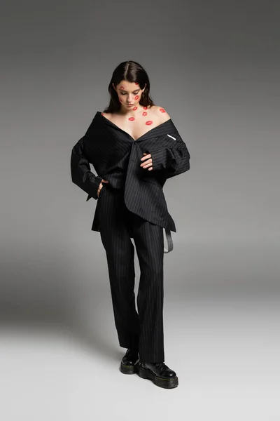 Full length of woman with red lip prints on bare shoulders and face standing in black oversize suit and rough boots on grey — Stockfoto