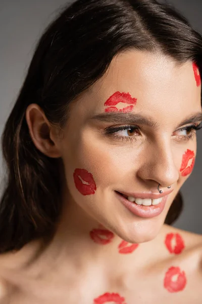 Portrait of happy woman with piercing and red lipstick marks on face looking away isolated on grey — Stock Photo