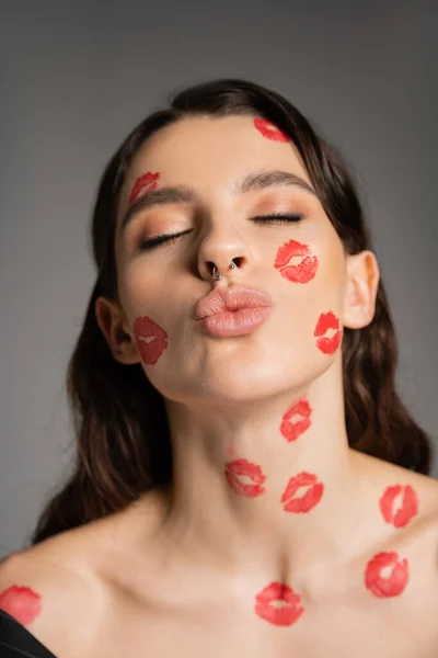 Portrait of sexy woman with closed eyes and red kiss prints on face pouting lips isolated on grey — Stockfoto