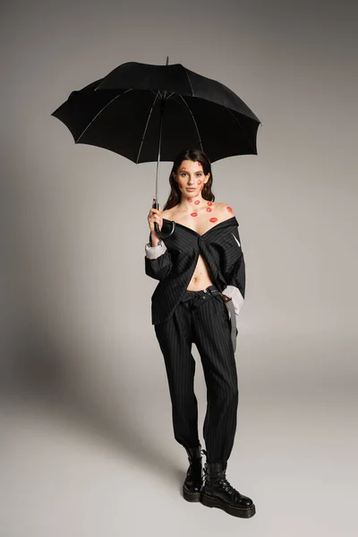 Full length of sexy woman with red kiss prints wearing oversize suit while holding umbrella on grey background — Stock Photo