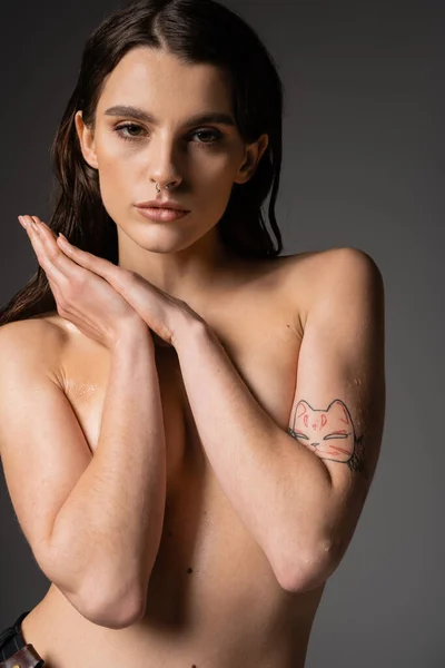 Sexy tattooed woman with shirtless body hiding breast with arms and looking at camera on grey background — Fotografia de Stock