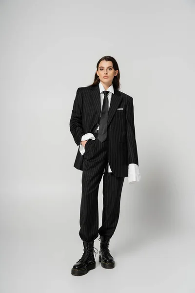 Full length of brunette woman in stylish formal wear and rough leather boots posing with hand in pocket on grey — Foto stock