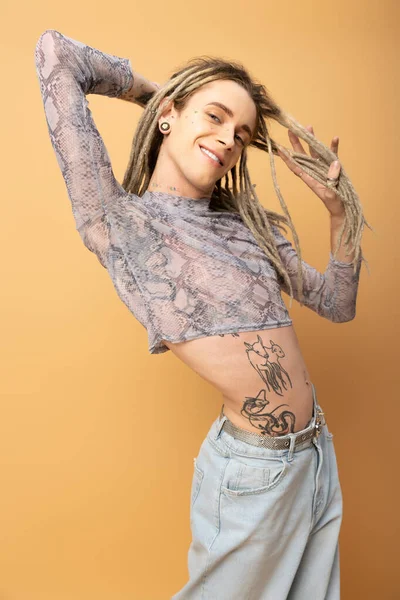 Smiling queer person touching dreadlocks and looking at camera isolated on yellow - foto de stock