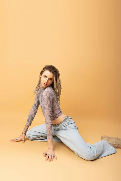 Stylish queer person in jeans and crop top sitting on yellow background — Photo de stock
