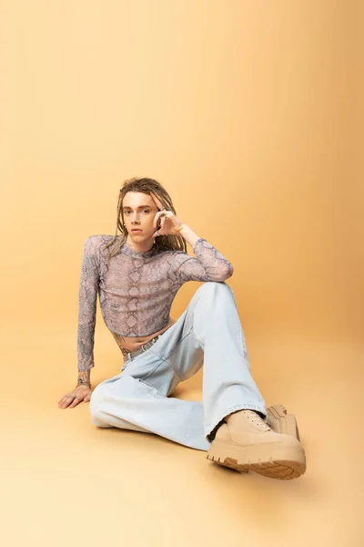 Full length of nonbinary person looking at camera while sitting on yellow background - foto de stock