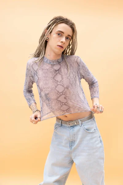 Young queer person with dreadlocks touching crop top with snakeskin print isolated on yellow — Stockfoto