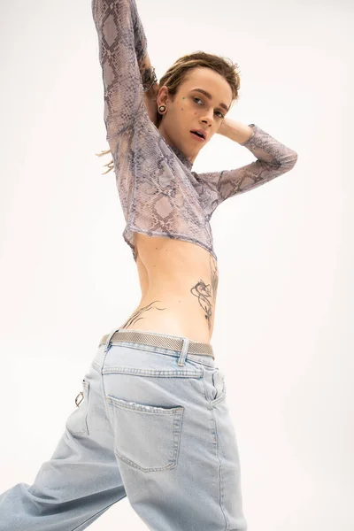 Low angle view of tattooed queer person in crop top with animal print looking at camera isolated on white - foto de stock