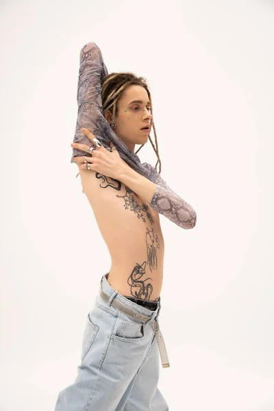 Tattooed queer person in jeans touching crop top isolated on white — стоковое фото