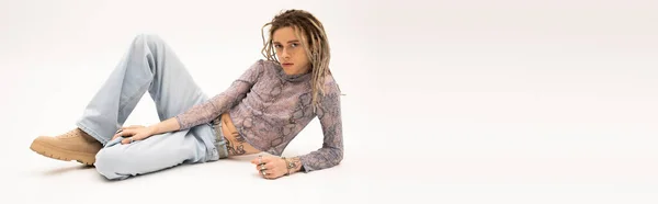 Queer person with dreadlocks looking at camera while lying on white background, banner — Stock Photo