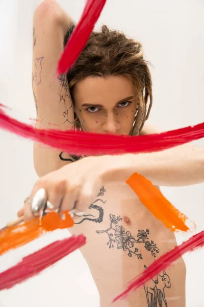 Tattooed nonbinary model looking at camera near blurred painted glass on white background - foto de stock