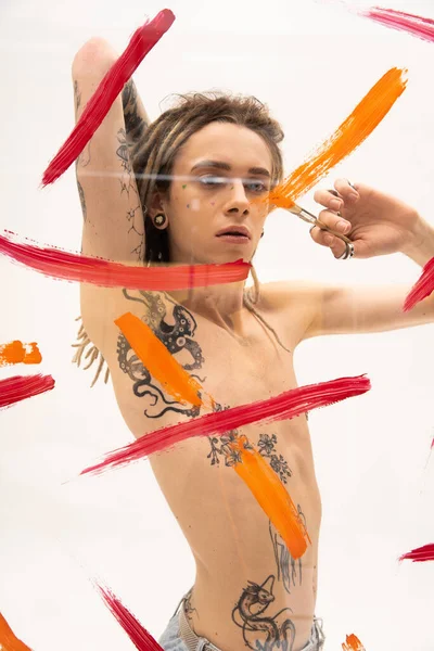 Shirtless nonbinary model with tattooed body painting on glass surface while standing on white background - foto de stock