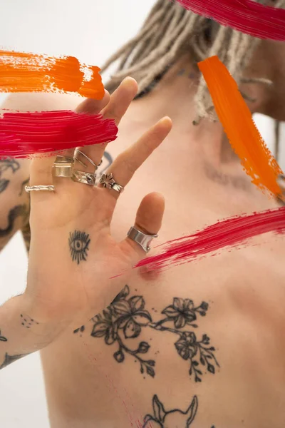 Cropped view of tattooed queer person touching glass with colorful paint strokes on white background — Stockfoto