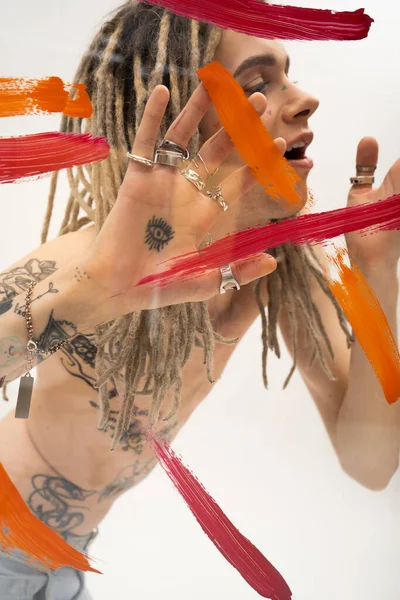 Tattooed nonbinary person in silver rings touching glass with multicolored paint strokes on white background — Stockfoto