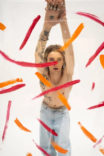 Shirtless and tattooed queer person in jeans standing with raised hands near glass with colorful strokes on white background — стоковое фото