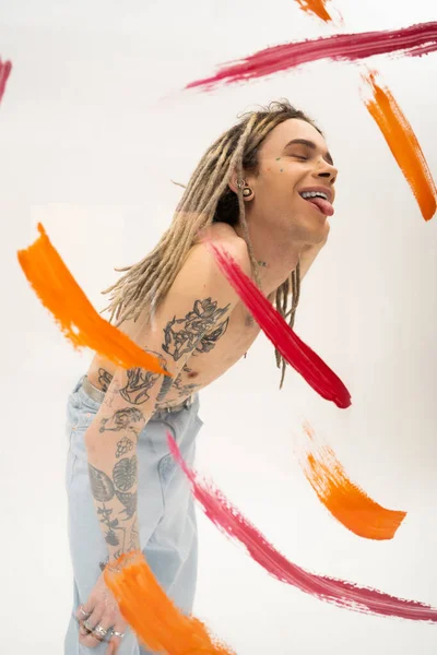 Positive nonbinary person with tattooed body sticking out tongue near colorful paint spills on white background — Stock Photo
