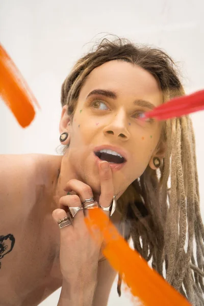 Retrato de modelo queer excitado con rastas tocando el labio y mirando hacia otro lado cerca de pinceladas en blanco - foto de stock