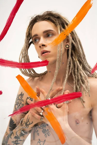 Young queer person with dreadlocks and tattooed body looking away near colorful paint spills on white background — стоковое фото