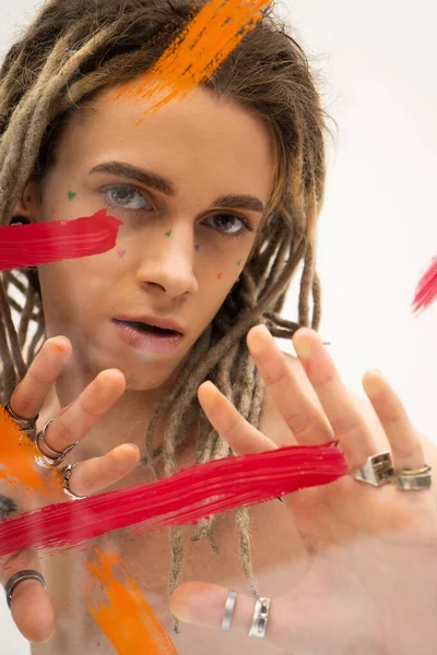 Portrait of queer model with dreadlocks and silver rings looking at camera near paint strokes on white background — Stock Photo