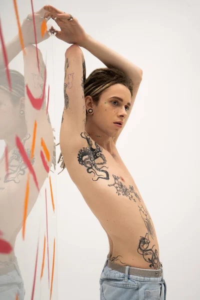 Young nonbinary model with tattooed body posing with raised hands near glass with paint spills on white background — Photo de stock