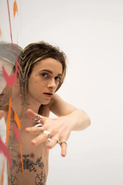 Young nonbinary person with silver rings on blurred hand looking at camera near glass with colorful strokes on white — Stockfoto