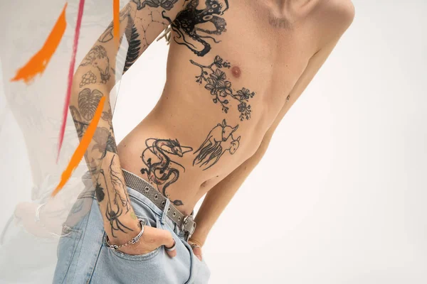 Cropped view of queer model with tattooed body posing with hands in pockets of jeans near glass with paint strokes on white background — Stockfoto