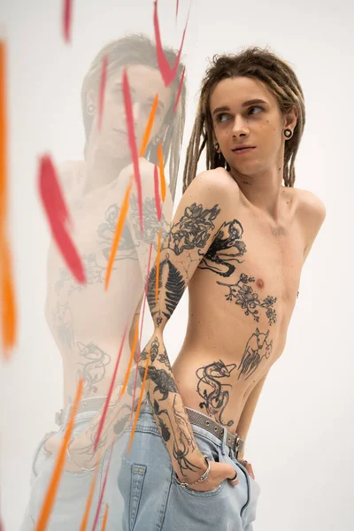 Young tattooed nonbinary model standing with hands in pockets of jeans near glass with paint strokes on white background — Stockfoto