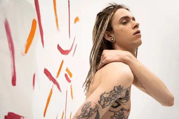 Side view of shirtless and tattooed queer model posing with hand on shoulder near multicolored brush strokes on white background — Stockfoto