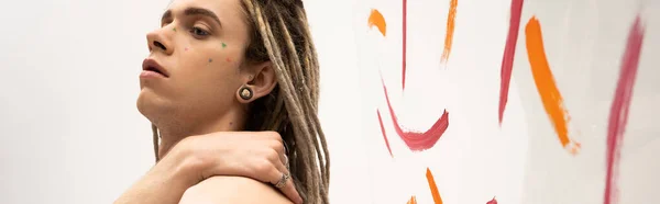 Portrait of queer person with dreadlocks posing with hand on shoulder near colorful brush strokes on white, banner — Stockfoto