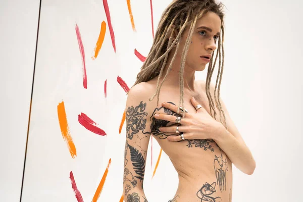 Безсорочка квір-людина з дредлоками, що торкаються татуйованого тулуба біля різнокольорових мазків фарби на білому тлі — стокове фото