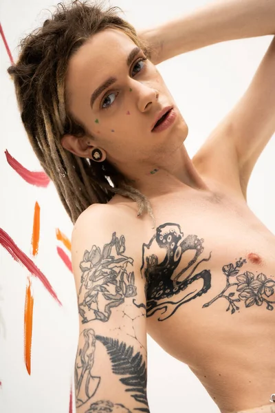 Young tattooed nonbinary model with dreadlocks looking at camera near colorful paint strokes on white — Stockfoto