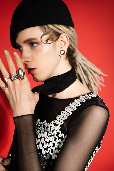 Nonbinary model with dreadlocks and silver finger rings holding hands near face and looking away on red - foto de stock