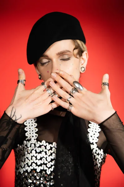 Queer personne avec les yeux fermés couvrant la bouche avec les mains en anneaux d'argent sur fond rouge — Photo de stock