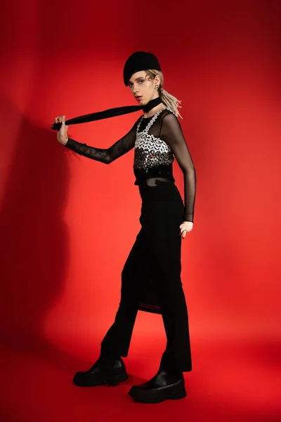 Full length of fashionable queer person holding black scarf and looking at camera on red with shadow — Stockfoto
