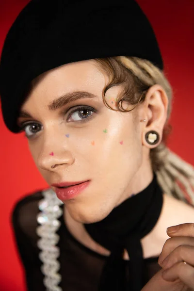 Close up portrait of tattooed queer person with makeup looking at camera on red background - foto de stock
