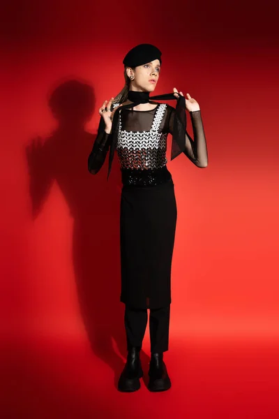 Full length of nonbinary model in beret and elegant top tying neckerchief on red background with shadow — Stock Photo