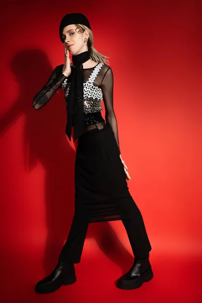 Full length of smiling nonbinary person in black elegant clothes holding hand near face on red background with shadow - foto de stock