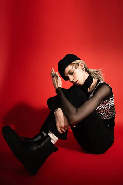 Full length of nonbinary model in black boots and beret sitting with closed eyes on red background — Stock Photo