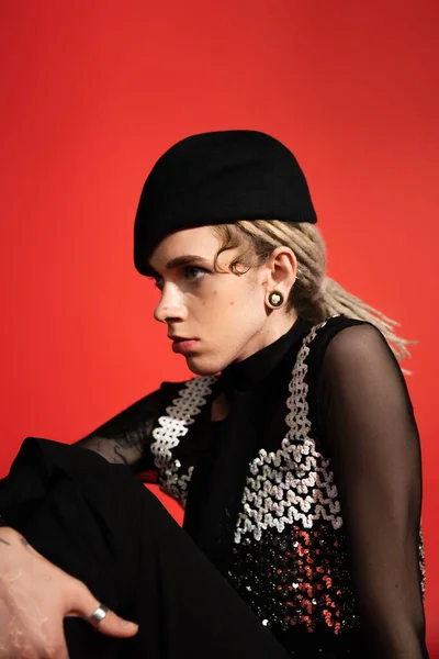 Stylish queer person in elegant clothes and black beret looking away while sitting isolated on red — Fotografia de Stock
