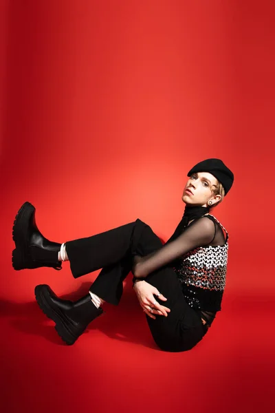 Full length of stylish queer model in black leather boots and beret sitting and looking at camera on red background — Stockfoto