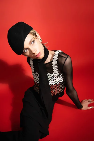 High angle view of trendy nonbinary person in black beret and top with sequins looking away on red background with shadow - foto de stock