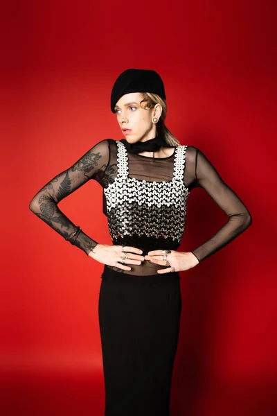 Tattooed queer model in black and elegant outfit posing with hands on waist and looking away on red background — Foto stock