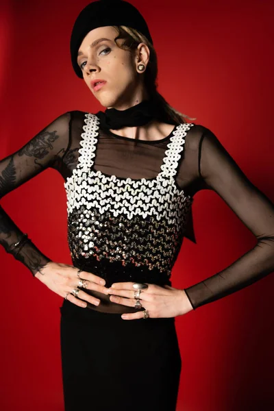 Tattooed nonbinary person in elegant clothes and black beret posing with hands on waist on red background - foto de stock