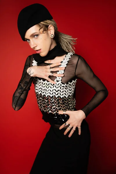 Elegant nonbinary model in black beret holding hand on chest while looking at camera on red background — Photo de stock
