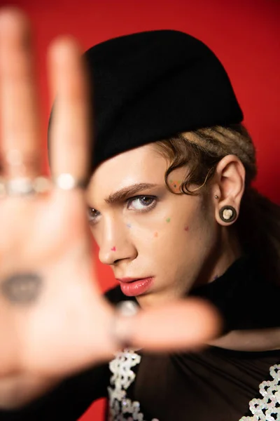Portrait of queer person in black beret looking at camera near blurred outstretched hand on red background — Stockfoto