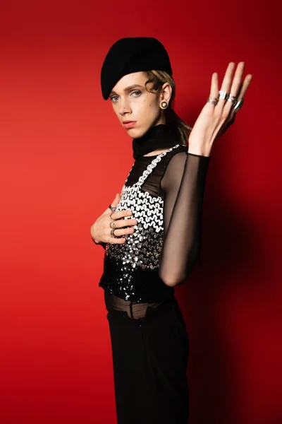 Stylish queer model in black beret and top with sequins pointing with hand and looking at camera on red background — Foto stock