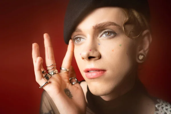 Portrait of queer model in black beret and silver rings touching face and looking away on dark red background - foto de stock