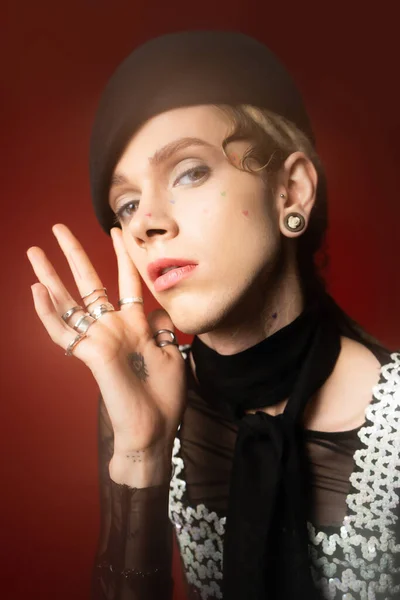 Portrait of young queer model in stylish attire and silver rings looking at camera on dark red background — Photo de stock